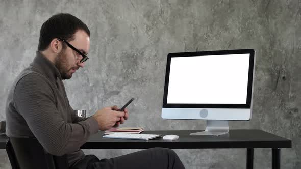 Businessman Looking at His Smartphone Messaging and Sitting Near Computer Screen. White Display alt