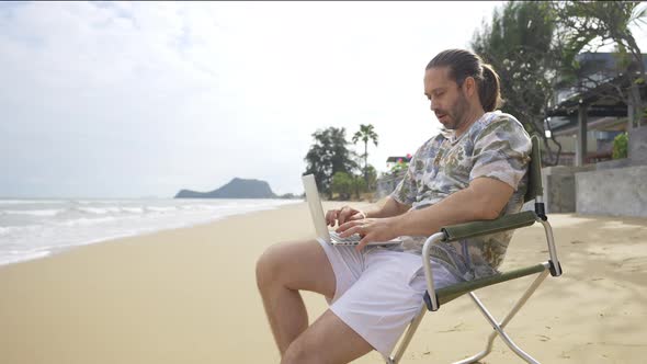 4K Caucasian businessman sitting on outdoor chair and working on laptop computer at the beach. alt