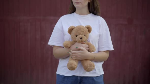 Unrecognizable Young Caucasian Woman Standing at Red Wall Outdoors Holding Teddy Bear alt