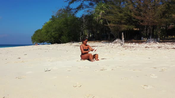 Woman spraying sunscreen on her tanned body, sitting on white sand of exotic beach, hot day of summe alt