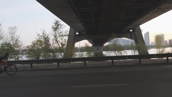 Silhouette of Young Sportive Cyclist Riding Road Bicycle in The City Under Huge Bridge at Sunset alt
