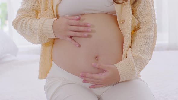 Close up Happy Pregnant Woman sitting on bed holding and stroking her big belly at home alt