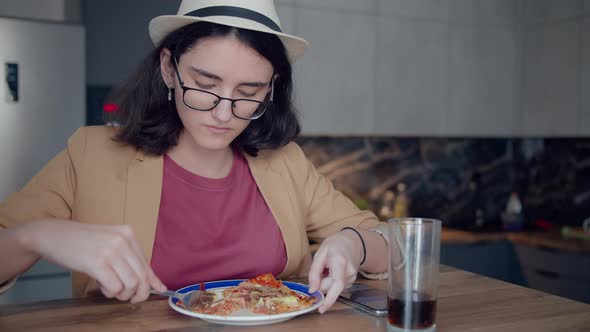 Pretty Teenage Girl in a Hat and Glasses Eating and Browsing a Mobile App alt