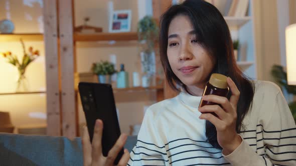 Sick young Asian lady hold medicine sit on couch video call with phone consult with doctor at home.