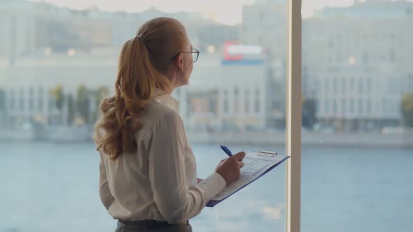 Attractive Smiling Mature Woman Standing at Window Indoors Writing Data in Clipboard alt