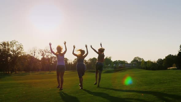 A Group of Carefree Young Women Running Along a Green Meadow, Waving Their Hands alt