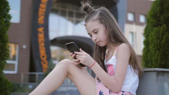 A Little Elementary School Girl Uses a Smartphone in the Background of a School or College Building