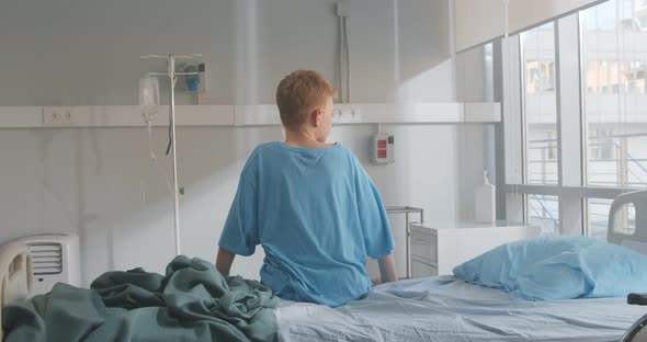 Back View of Kid Patient Sitting on Hospital Bed Looking Out of Window alt