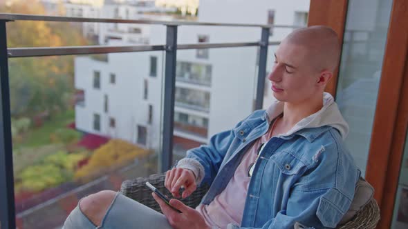 Confident Bald Queer Gay Guy Sitting on an Armchair on His Balcony Looking Both Sides Relaxing Using alt