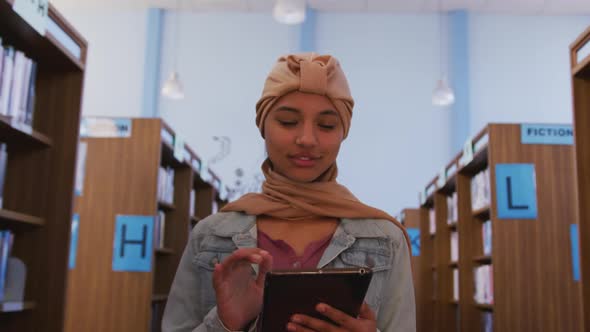 Asian female student wearing a beige hijab walking and using a tablet alt