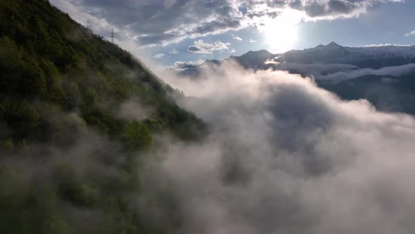 Flight above gray puffy clouds enveloping dark forested alp hillside in sunlight alt