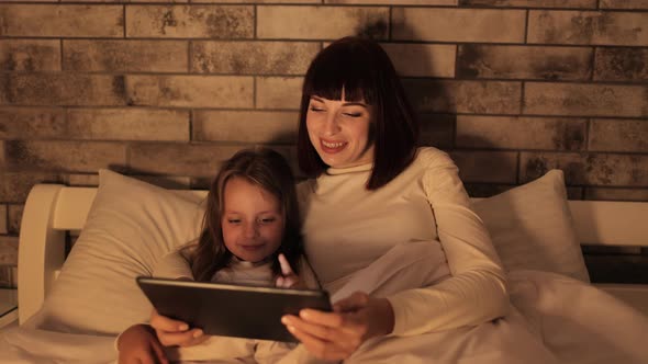 Mother and Little Preschool Daughter Lying Under Blanket on Bed Watching Video on Tablet alt