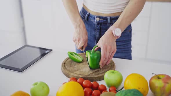Young female is cutting green pepper on a wooden board. alt