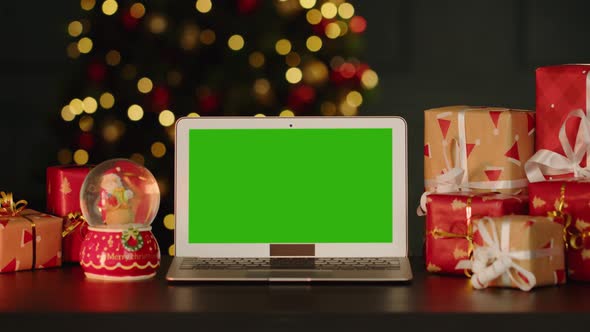 Laptop with Green Screen on Table with Christmas Decorations Against Christmas Tree