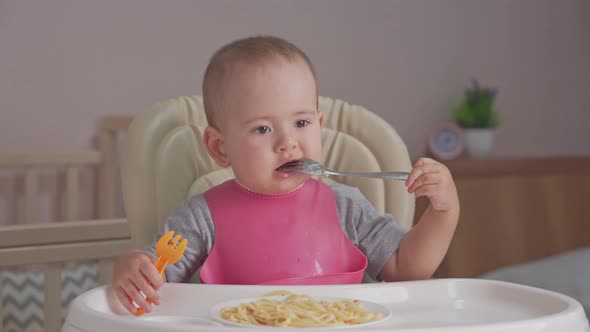 Toddler eats spaghetti for dinner by himself holding two forks in his hands alt