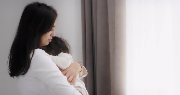 Mother Near the Window in the Bedroom Raises Her Baby Daughter in Her Arms