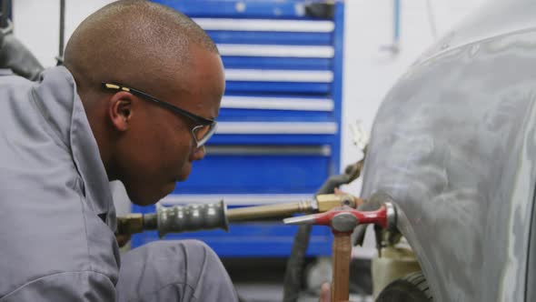 African American male car mechanic holding a screwdriver and using a hammer to repair a car alt
