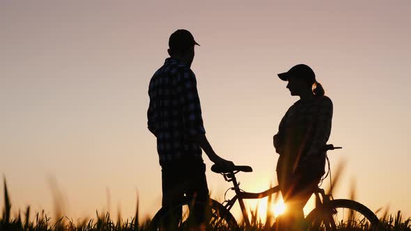A Man and a Woman Enjoy an Evening Walk at Sunset Standing with a Bicycle in Nature alt