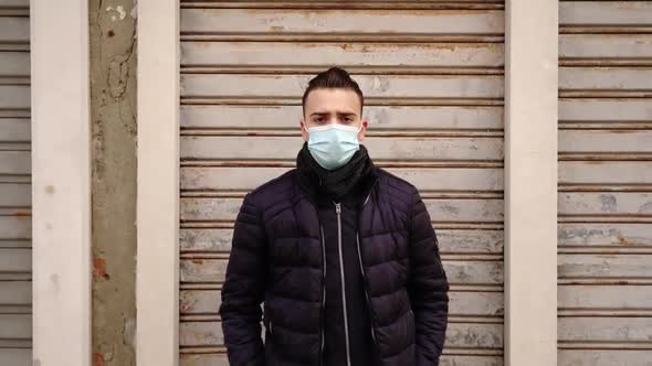 Man with Medical Mask Stands Near Light Grey Closed Shutters alt