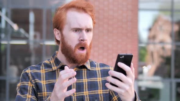 Outdoor Redhead Beard Young Man Upset By Loss While Using Smartphone alt