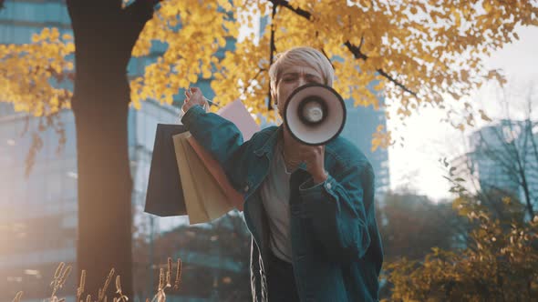 Young Blond Woman with Shopping Bags Announcing Sales Season with Loudpeaker in the City Park in alt