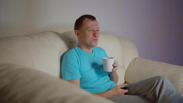 Lonely young man with a mug of tea sits on the sofa in the evening and watches TV