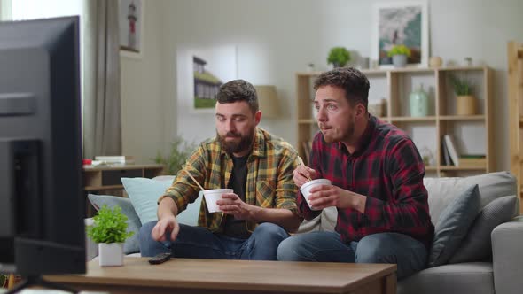 Two Brothers Dine Noodles From Delivery and Watch News Sitting on Sofa in the Living Room alt