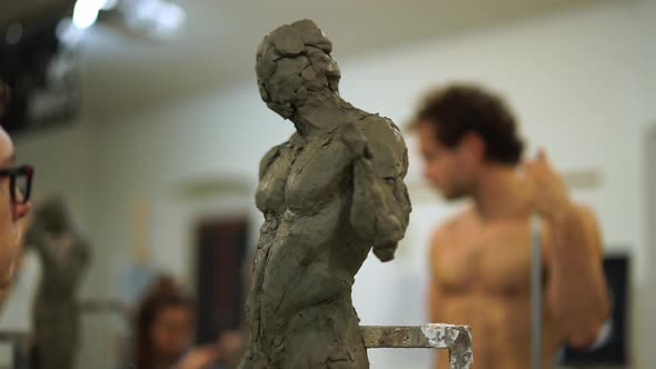 Student working on clay sculpture, nude model in background