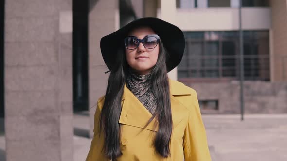 Stylish Girl with a Good Mood in a Yellow Raincoat Hat and Glasses Comes with Luggage alt