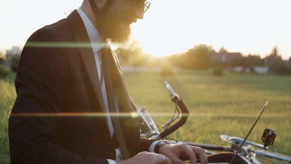 Man Sitting on a Grass at the Park Working on a Laptop Wearing a Helmet alt