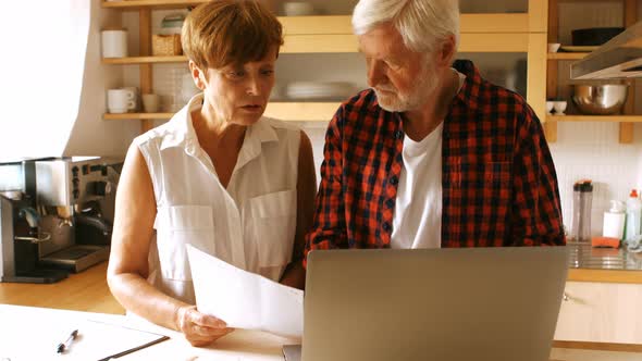 Senior couple paying bills online on laptop in kitchen alt