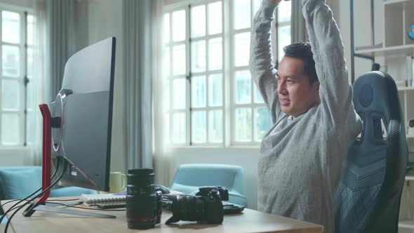 Asian Cameraman Stretching And Leaning Back While Using Desktop Computer For Working At Home alt