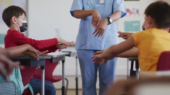 Diverse schoolchildren and medical worker wearing face masks, disinfecting hands in classroom alt