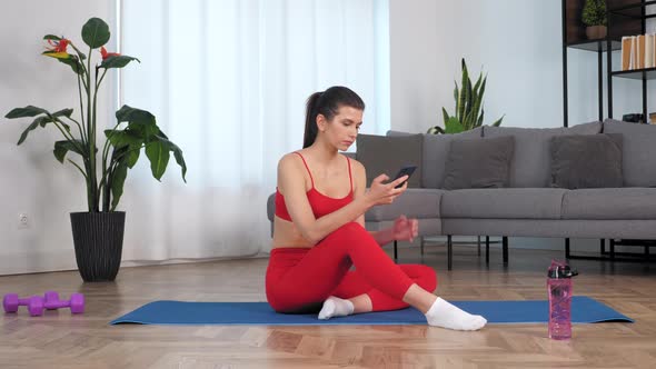 Sportive Beautiful Woman in Sportswear Sitting on Yoga Blue Mat Uses Smartphone alt
