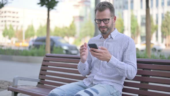 Young Adult Man Celebrating Online Success on Smartphone While Sitting Outdoor on Bench alt