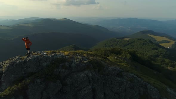 Young Man in Orange Jacket Standing at the Edge and Enjoys Success Achieved From Heavy Climbing alt