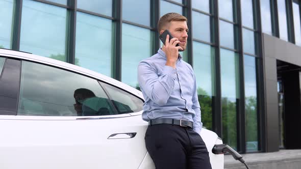 Portrait of Young Handsome European Man Speaking on His Smartphone While Leaning on His Electric Car alt