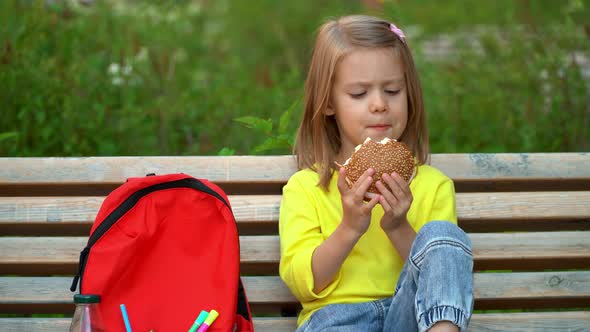Little Schoolchild Sits Outdoors and Eats Hamburger