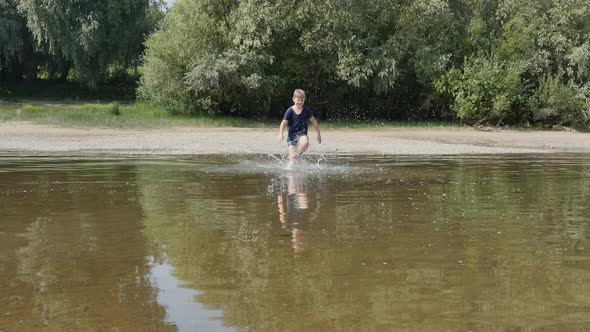 Caucasoid Teen Boy in a Tshirt and Shorts Runs Along the River Splashing Water
