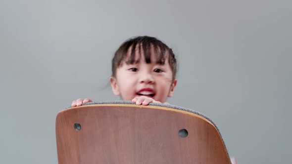 Girl playing to hide behind wooden armchair. Engraved in studio with gray background. alt