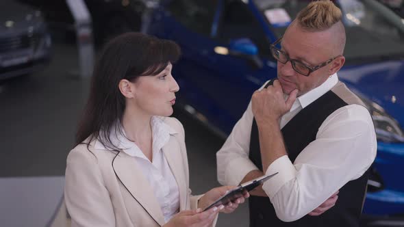Thoughtful Caucasian Man in Eyeglasses Listening Woman Talking Explaining Vehicle Advantages in Car alt