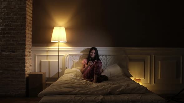 Beautiful girl in pink pajamas sitting in the evening on the bed with the phone alt