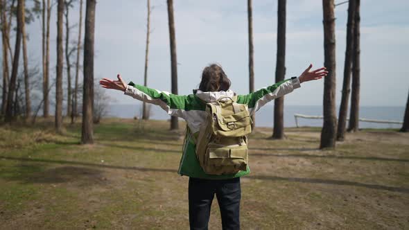 Back View Portrait of Excited Traveler Stretching Hands Standing on Forest Meadow in Front of River alt
