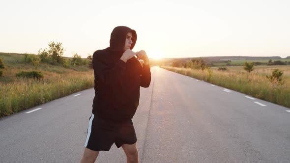 Kickboxer with Hood on Head Training Kicks on Country Road Singly During Sunset alt