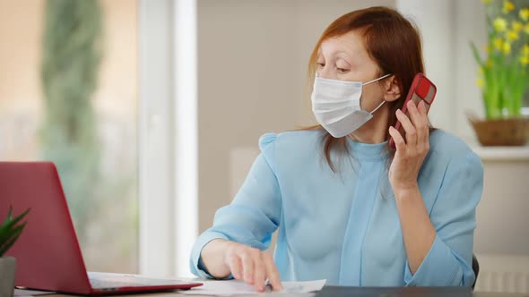 Portrait of Busy Woman in Coronavirus Face Mask Talking on Phone Messaging Online on Laptop alt