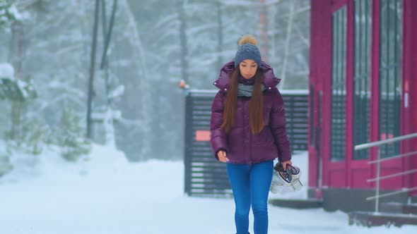 Woman in Purple Jacket Walks Holding Skates Along Snowy Park