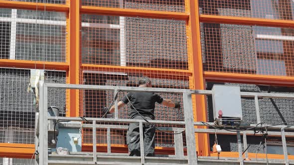 A Builder on a Lift Installs a Protective Metal Mesh on the Facade of the House alt