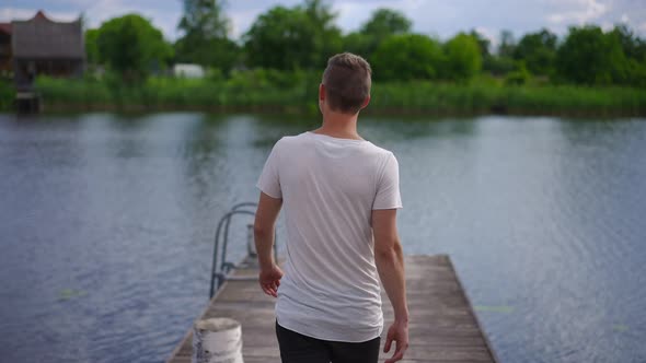 Back View Confident Young Caucasian Man Walking in Slow Motion to River on Wooden Pier Raising Arms alt