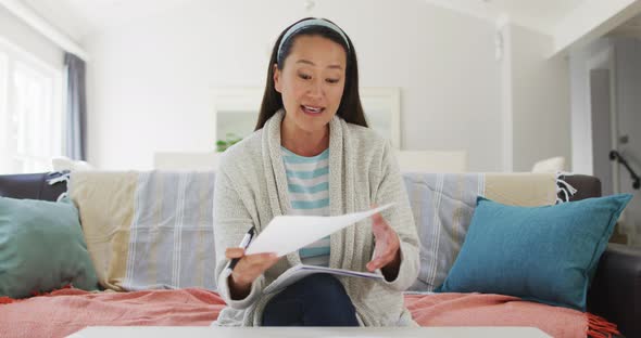 Asian woman working at home, sitting on couch making video call, talking and reading paperwork alt