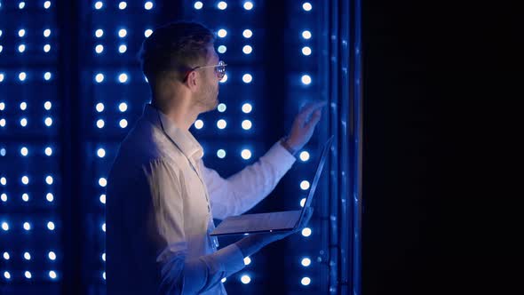 Male Network Engineer Doing a System Check Standing in the Server Room with His Laptop alt